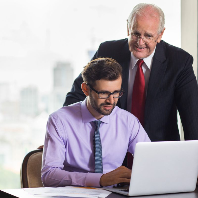 Senior businessman standing behind young colleague sitting at table with big window and blurry city view in background. They are looking at laptop screen.